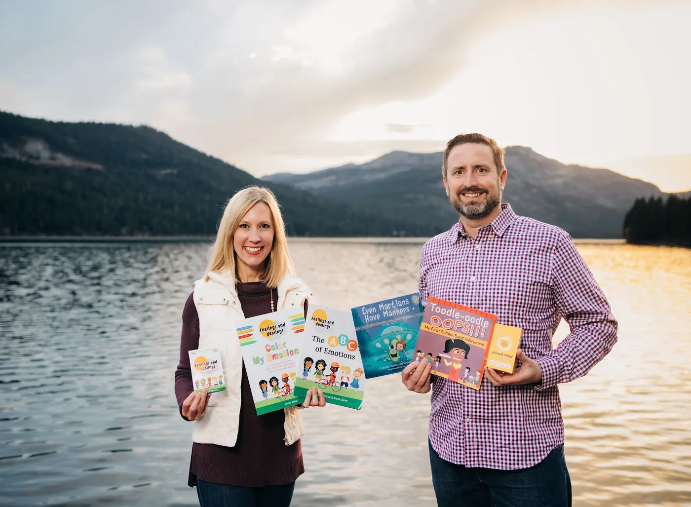 Cami Childs and Bryan Jones standing by Donner Lake holding the games and books they have made, including Feelings and Dealings, Color My Emotions, The ABCs of Emotions, Even Martians Have Manners and Toodle-oodle Oops